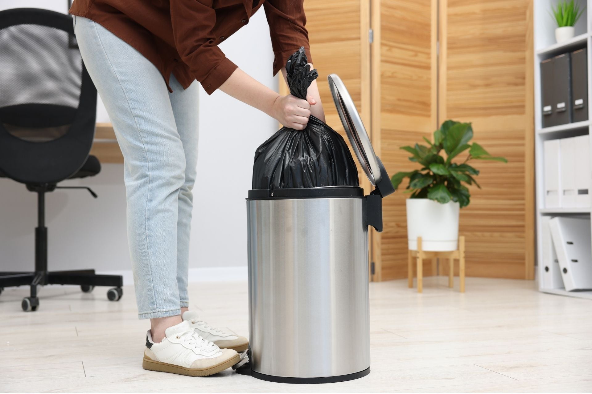 Person lifting a tied black trash bag out of a stainless steel step trash can indoors