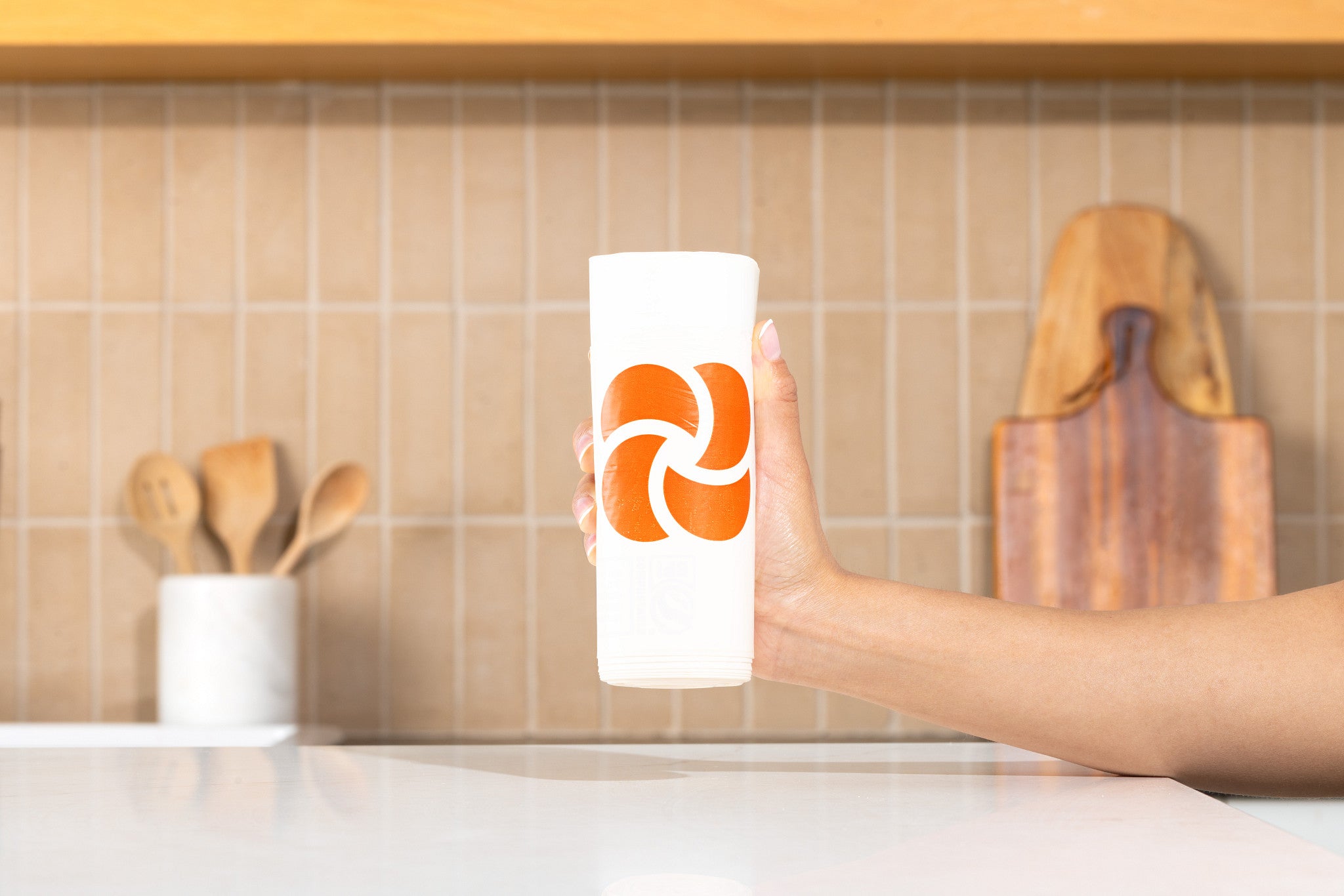 Hand holding a roll of Plastno compostable waste bags with the orange logo, in a kitchen with tile backsplash