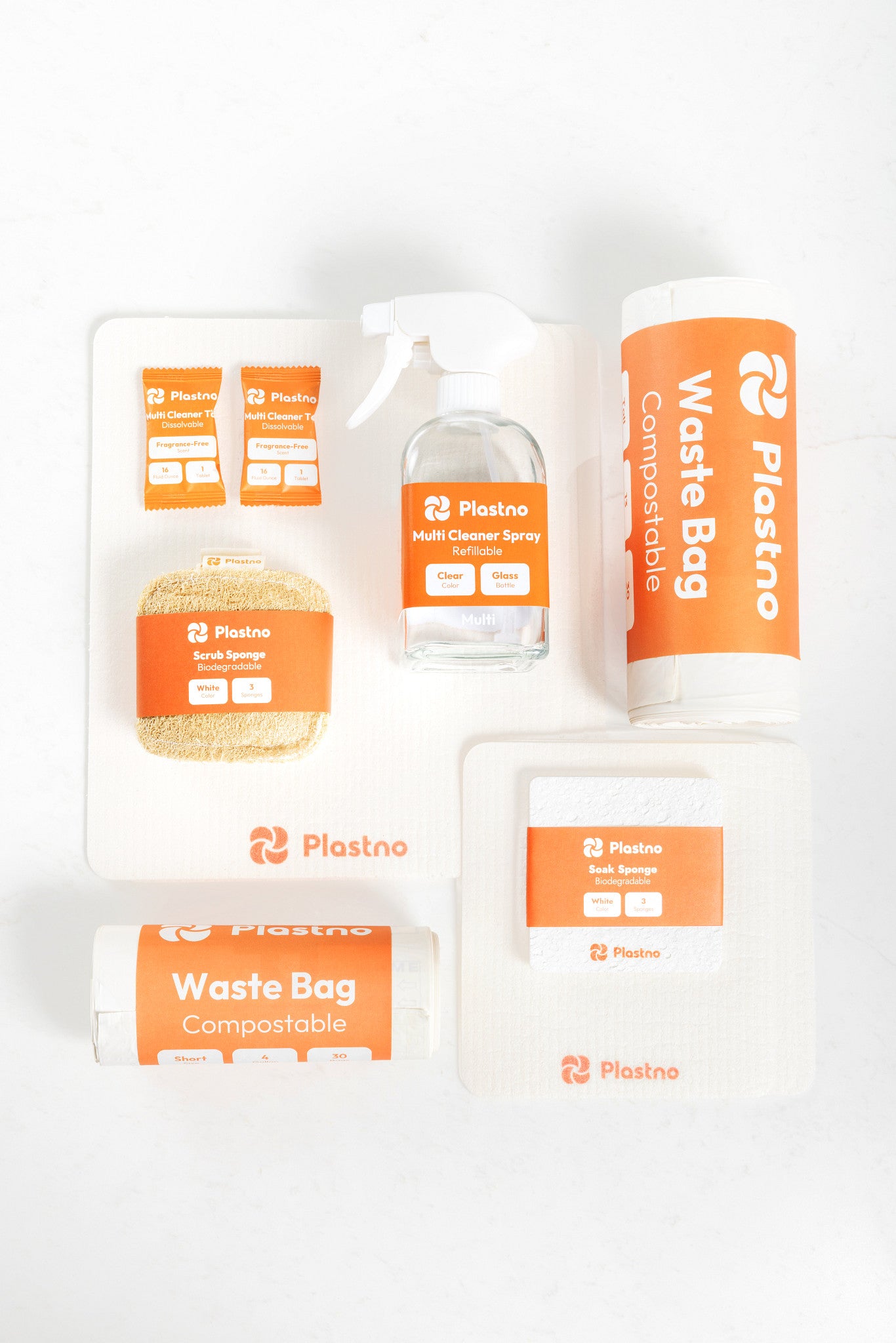 Flat lay of the Plastno cleaning bundle: multi cleaner tablets, refillable glass spray bottle, scrub sponge, soak sponge, sponge cloth and two rolls of compostable waste bags