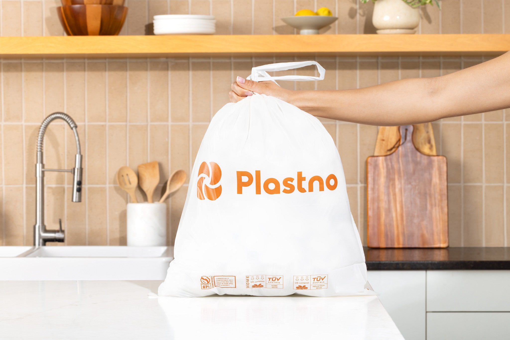 Hand lifting a full Plastno compostable waste bag by its drawstring handles on a kitchen counter, showing the orange logo and BPI and TUV compost certification marks