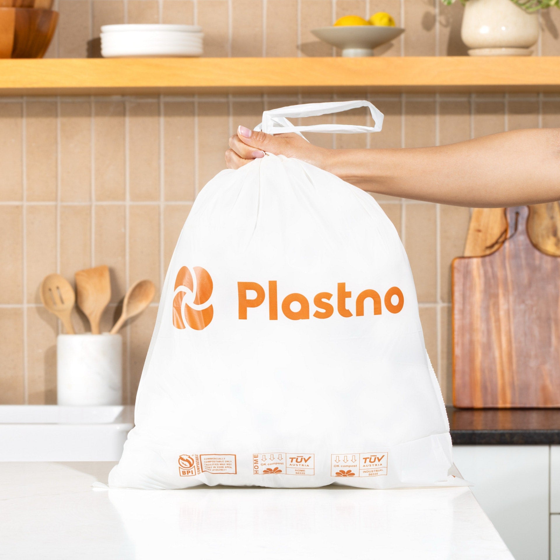 Plastno Waste Bag Short: hand lifting a 4 gallon compostable trash bag closed with its drawstring, showing the BPI and TUV compost certification marks