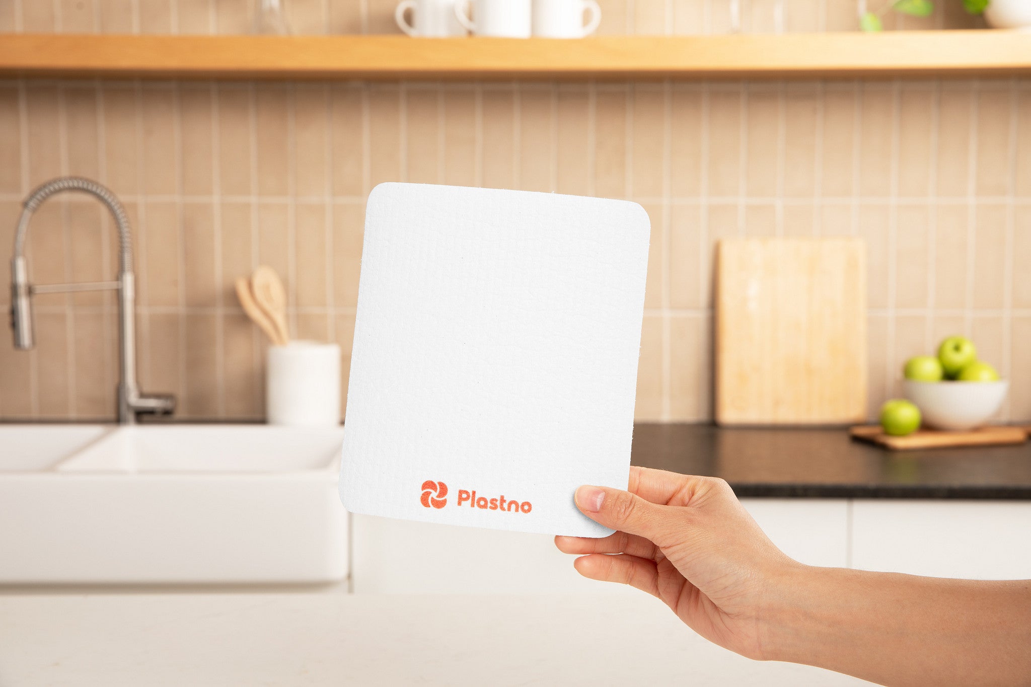 Hand holding a white Plastno sponge cloth with orange logo in front of a kitchen sink and tiled backsplash