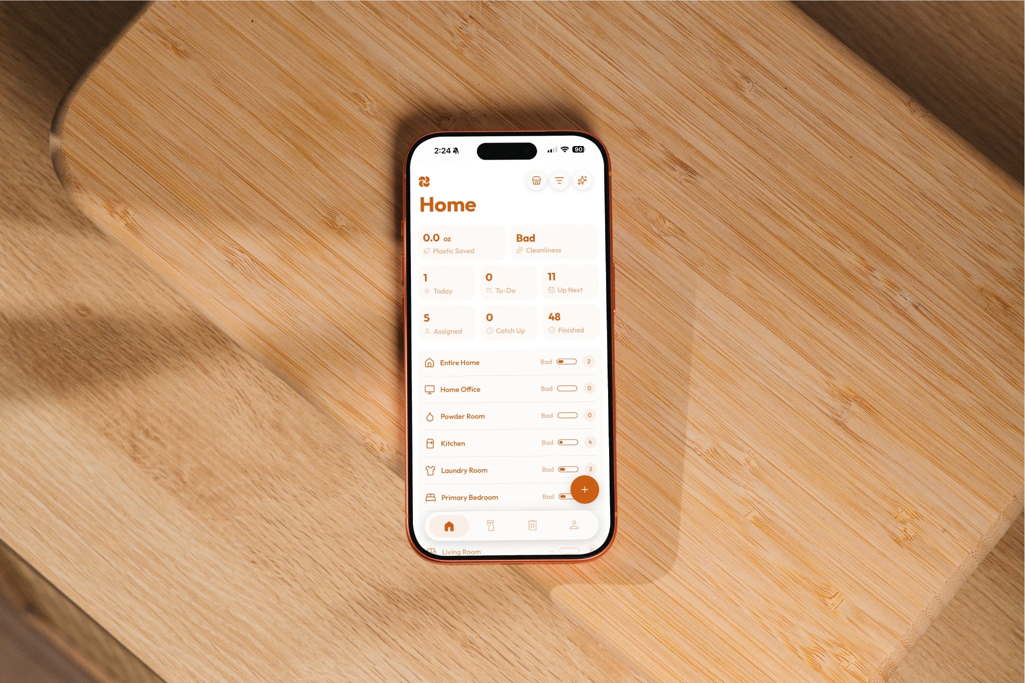 Plastnofy app home screen on an iPhone resting on a wooden board, showing plastic saved, cleanliness score, task counts and a room-by-room list