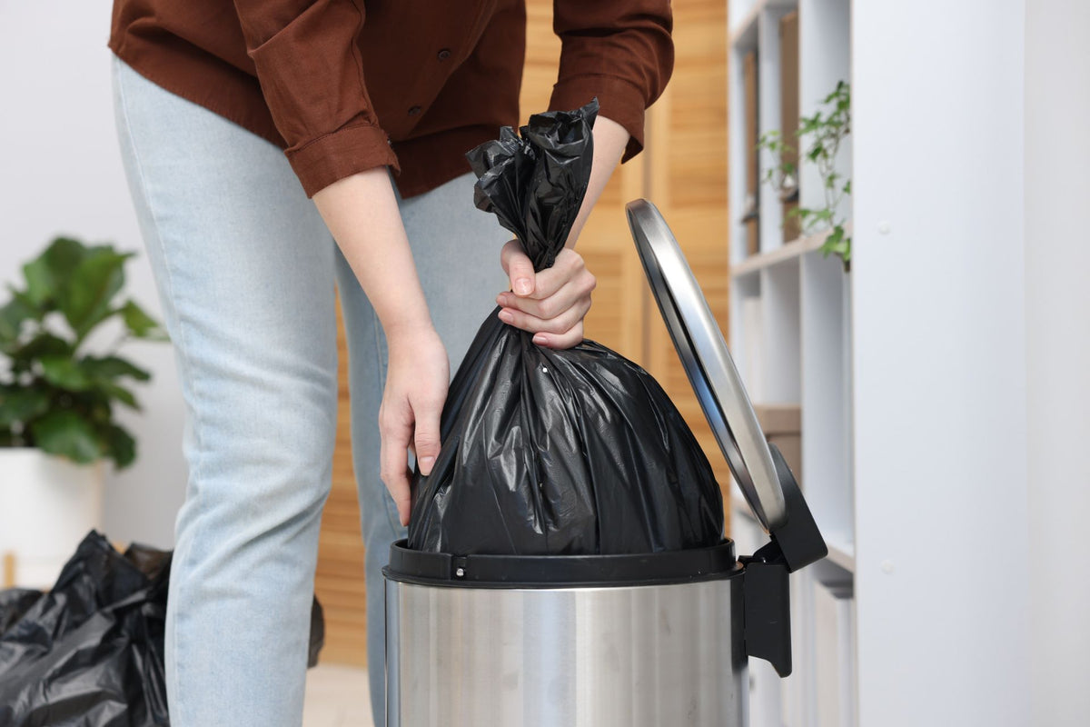 Person lifting a full, tied black trash bag out of a stainless steel step trash can