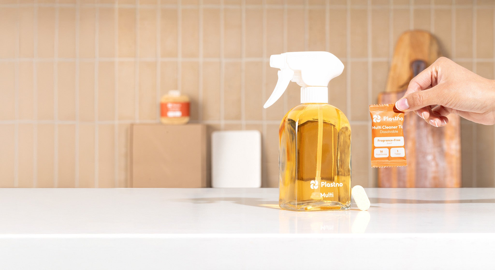 Plastno glass spray bottle filled with Multi cleaner beside a dissolvable multi cleaner tablet and a hand holding its fragrance-free refill packet