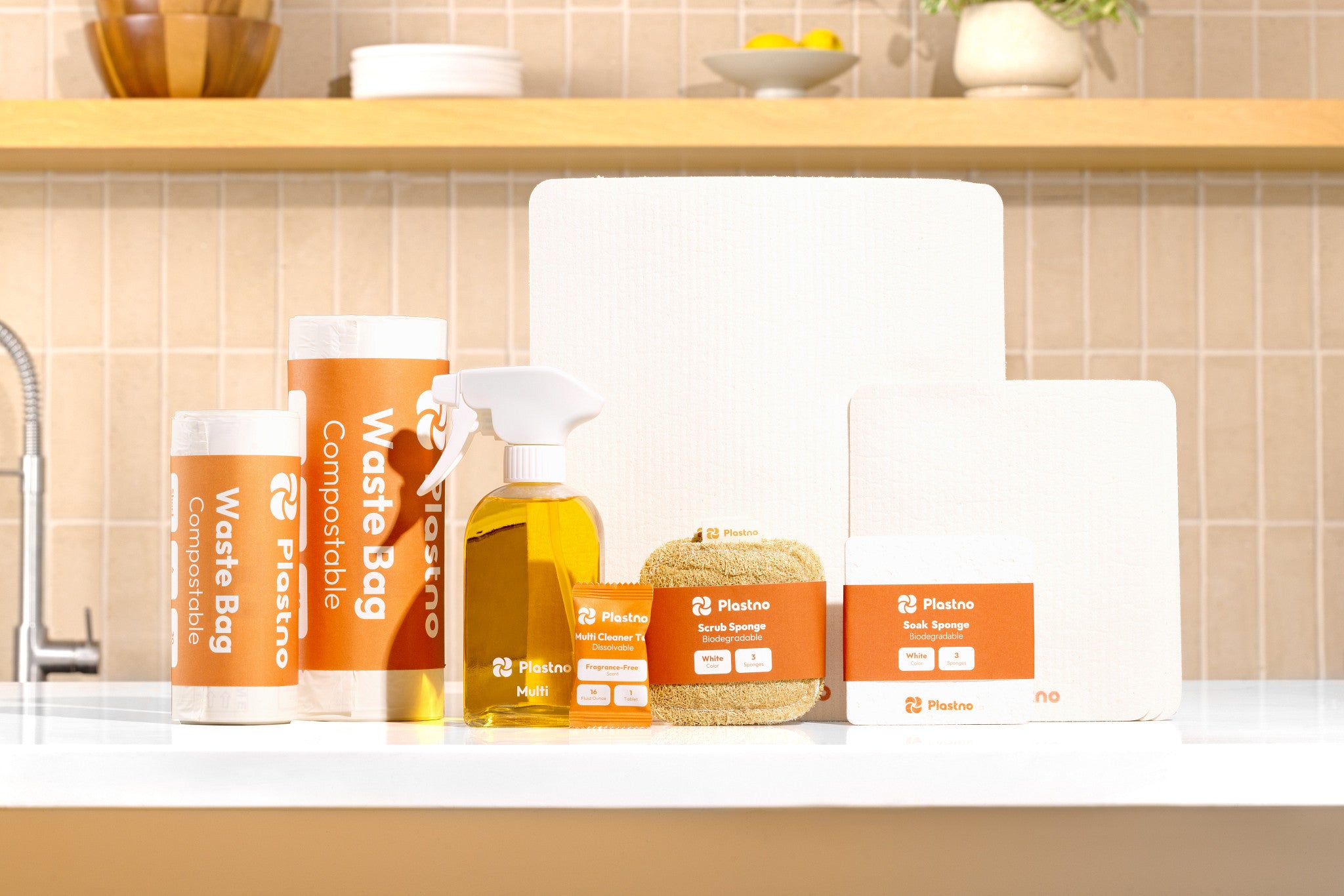 Full Plastno cleaning bundle on a kitchen counter: two rolls of compostable waste bags, Multi cleaner glass spray bottle, cleaner tablet, scrub sponge, soak sponge and sponge towels