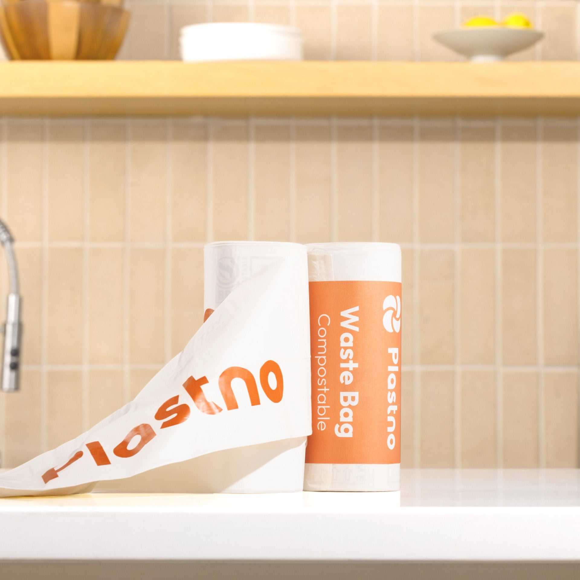 Two rolls of Plastno compostable waste bags on a white kitchen counter, one unrolled to show the orange Plastno logo, with a tiled backsplash in the background.