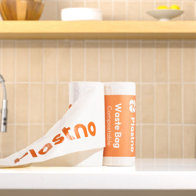 Two rolls of Plastno compostable waste bags on a white kitchen counter, one unrolled to show the orange Plastno logo, with a tiled backsplash in the background.