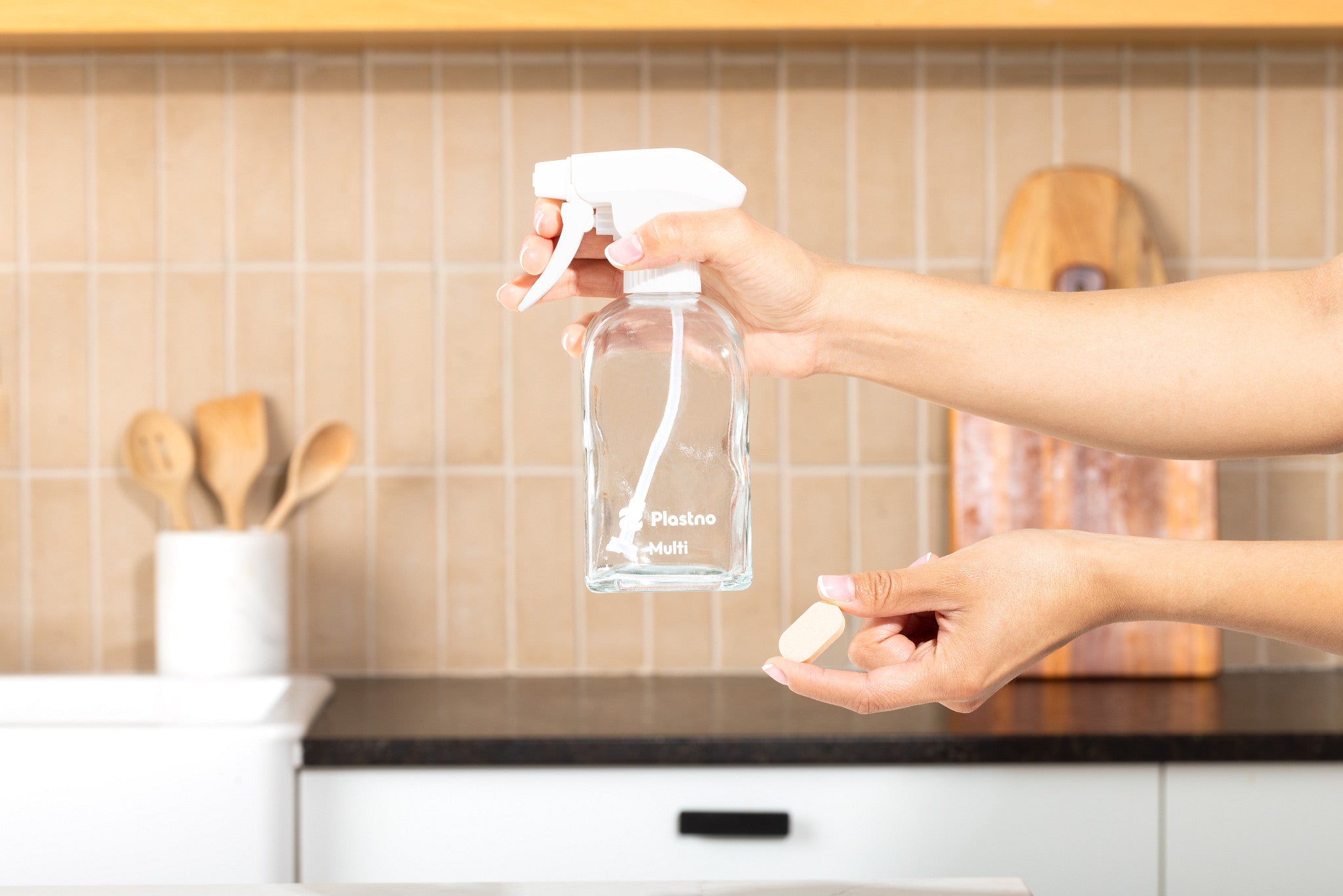 One hand holding an empty Plastno Multi glass spray bottle and the other holding a dissolvable cleaner tablet in a kitchen
