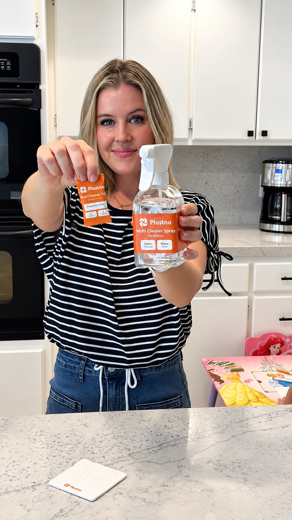 Person in a kitchen holding up a Plastno Multi Cleaner Tablet packet in one hand and the refillable Multi Cleaner Spray glass bottle in the other, with a Plastno sponge cloth on the counter