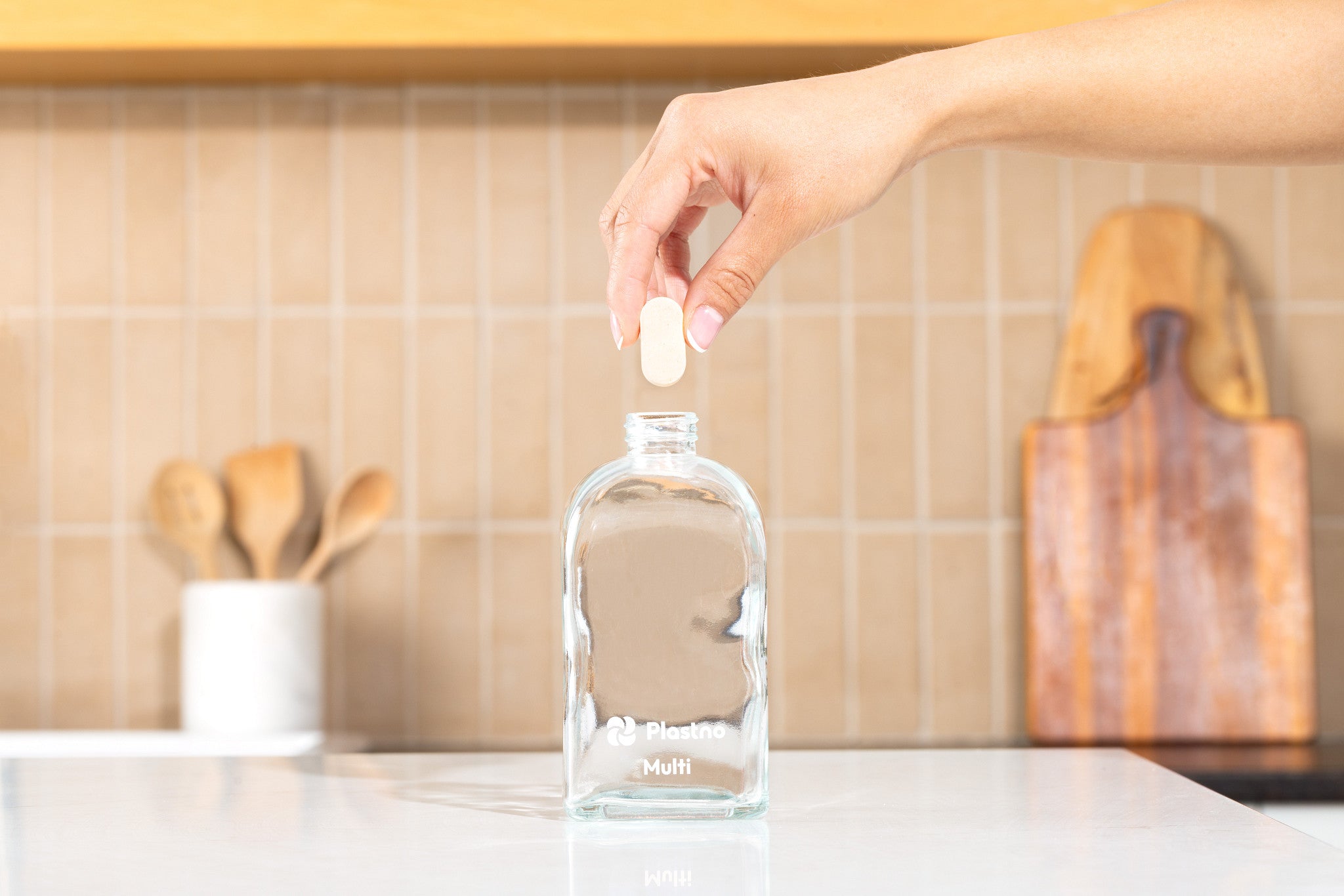 Hand dropping a Plastno multi cleaner tablet into an empty Plastno Multi glass bottle on a kitchen counter