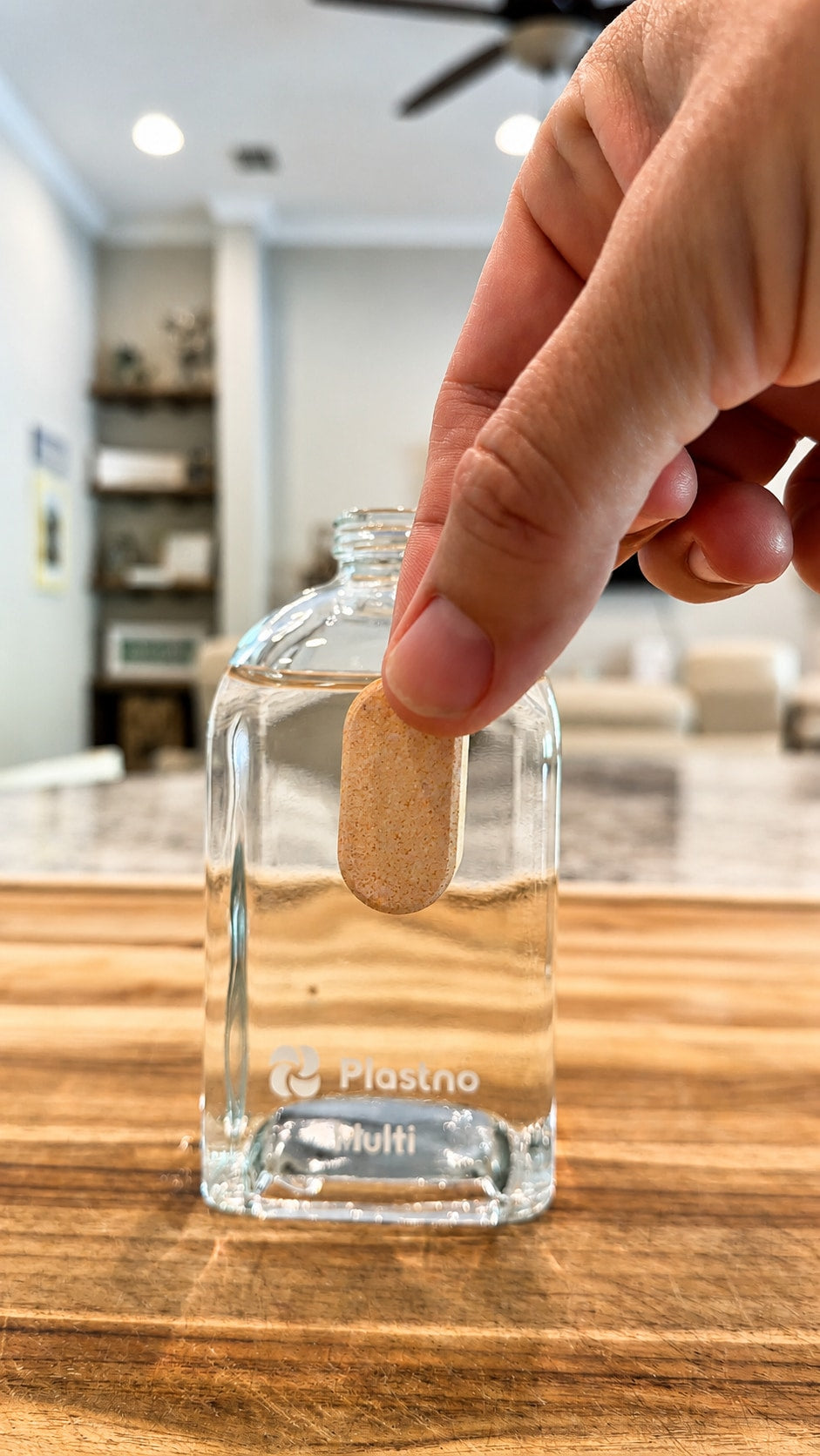 Hand lowering a Plastno multi cleaner tablet into a water-filled Plastno Multi glass bottle on a wooden counter