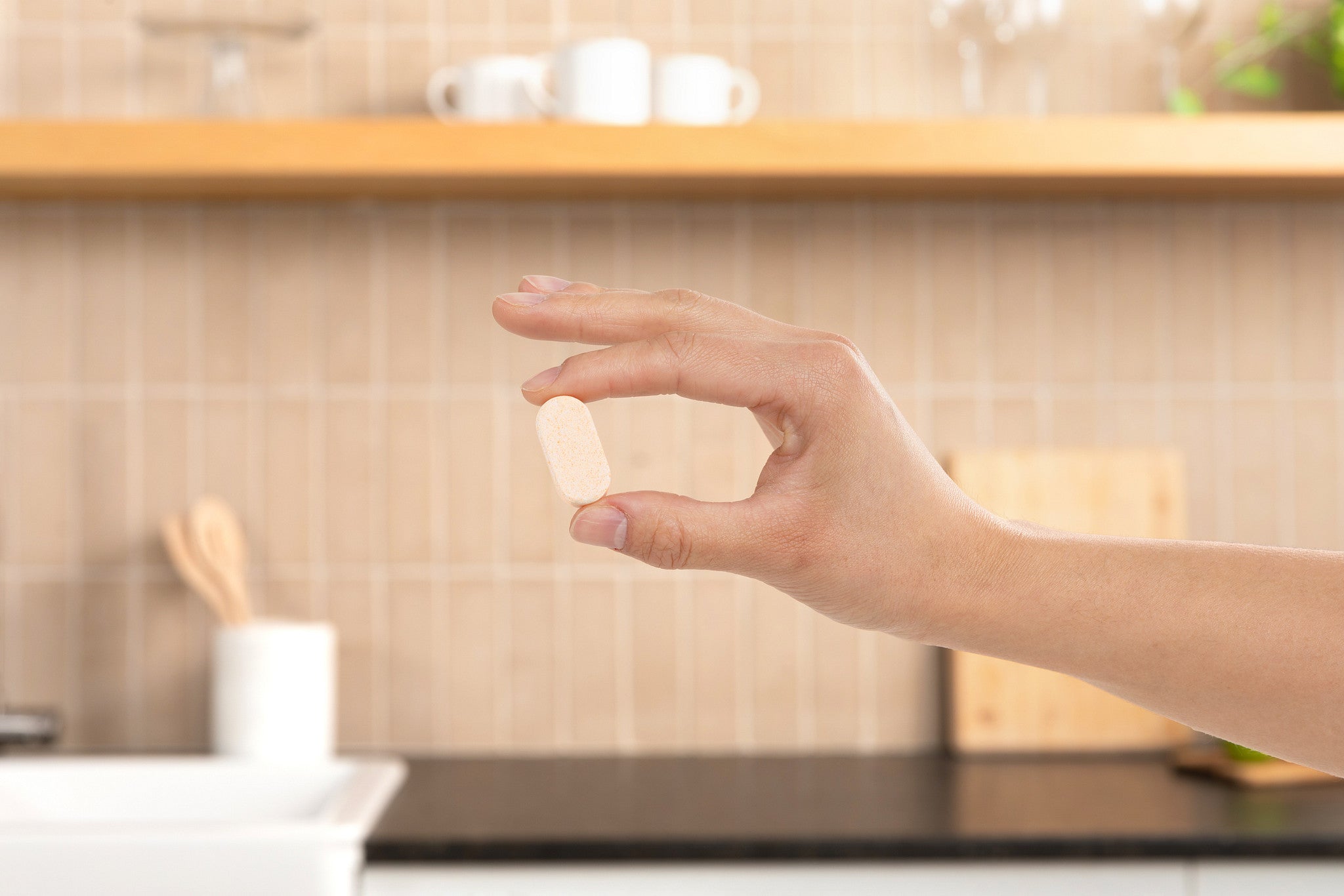Hand holding a single Plastno dissolvable multi cleaner tablet in front of a tiled kitchen backsplash