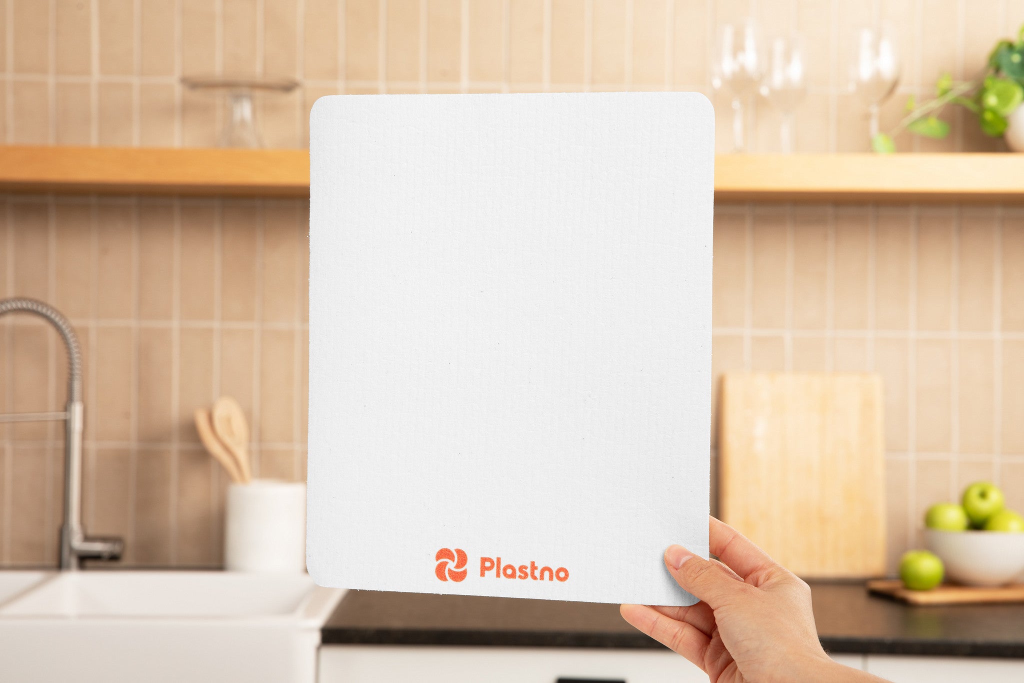 Hand holding a large white Plastno sponge towel with orange logo in front of a kitchen sink and tiled backsplash