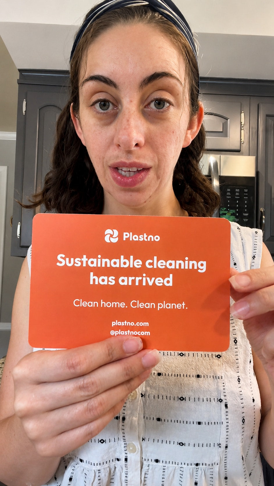 Person in a kitchen holding an orange Plastno card that reads "Sustainable cleaning has arrived, Clean home. Clean planet."